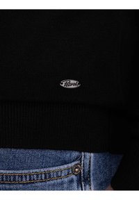 Primo piano di un cardigan nero in maglia con il logo argentato "Petrol" sopra la tasca dei jeans blu e il bottone con il testo del marchio.