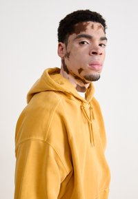 Sweatshirt à capuche en coton jaune avec cordons de serrage, présentant une coupe décontractée et une texture subtile. Le modèle a des marques de peau visibles.