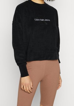 Person iført en sort Calvin Klein Jeans sweatshirt og brune tætsiddende knælange shorts mod en ensfarvet baggrund.