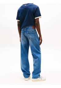 Personne portant une chemise bleue à manches courtes et un jean bleu à coupe décontractée, debout les mains le long du corps, de dos sur fond blanc.