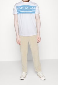 T-shirt blanche à manches courtes avec logo bleu "TOM TAILOR" et rayures, associée à un pantalon en velours côtelé beige clair et des baskets blanches.