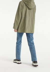 Oliwkowa kurtka z kapturem o oversize'owym kroju, w zestawieniu z jasnoniebieskimi dżinsami i wielokolorowymi sportowymi butami.