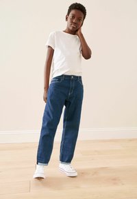 Jeans de denim azul con un corte relajado y bolsillos frontales, combinados con una camiseta básica blanca de cuello redondo y zapatillas blancas con detalles rojos.
