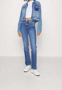 Modrá džínová bunda s kratším střihem a roztřepeným lemem, spárovaná s úzkými modrými džíny a bílými teniskami. Obsahuje klasické kapsy a švy.