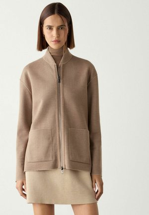 Ung kvinde med glat brunt hår iført en beige cardigan med lynlås over et matchende skørt og rullekrave, stående mod en ensfarvet baggrund.