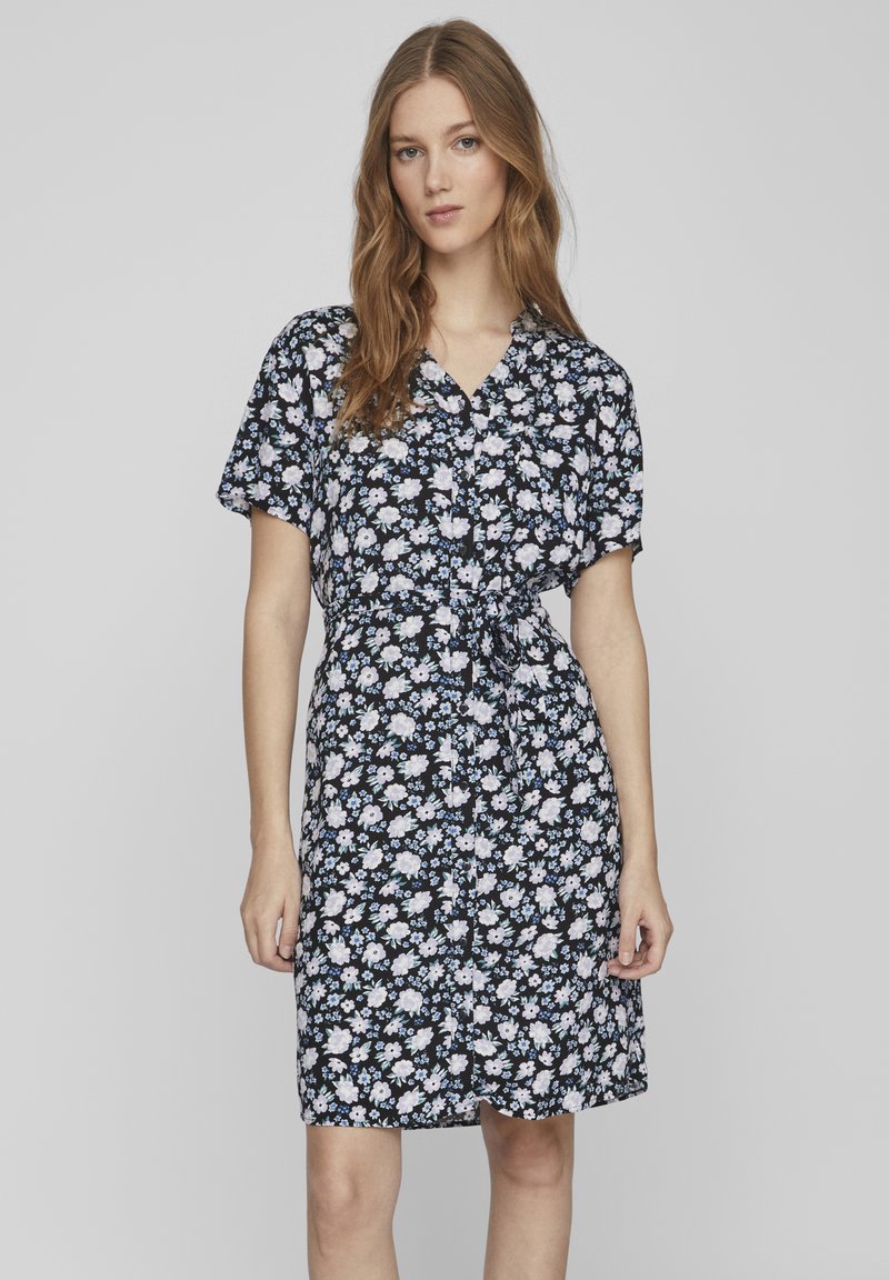 Abito floreale di un blu navy scuro, con maniche corte, apertura frontale con bottoni e cintura da annodare. Il tessuto presenta piccoli fiori bianchi e azzurri chiari.