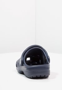 Granatowy gumowy klompk z regulowanym paskiem z tyłu i teksturowaną podeszwą, pokazany z tyłu na białej powierzchni i tle.