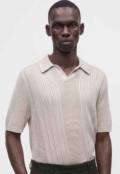 Man with dark skin wearing a light beige textured short-sleeve polo shirt standing against a plain light background.
