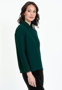 Garās piedurknes, apkakles krekls dziļi zaļā audumā ar gludu tekstūru, pielāgota piegriezuma un taisnu apakšmalu. Valkāts kopā ar melniem biksēm.