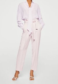 Lichte lavendelkleurige blouse met een strik, gecombineerd met hoogopgetrokken lichtroze broeken. Glad materiaal, relaxte pasvorm, geaccentueerd met een ceintuur in de taille. Witte hakken.