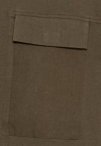 Tecido verde oliva com um bolso com remendo visível. O material apresenta uma textura suave e é costurado nas bordas para maior durabilidade.