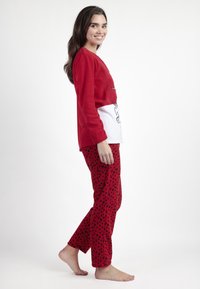 Joven mujer con una blusa de manga larga roja y blanca y pantalones rojos con lunares, de pie descalza y sonriendo frente a un fondo blanco liso.
