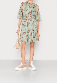 Blumendruck, mintgrünes Kleid mit halbtransparenten Ärmeln, elastischem Bund und Rüschensaum. Kombiniert mit beigen High-Top-Sneakers und einer kleinen Tasche.