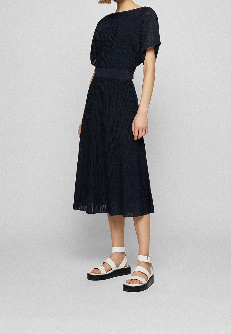 Femme portant une robe midi noire à manches courtes, taille cintrée, et des sandales blanches à plateforme sur un fond gris uni.