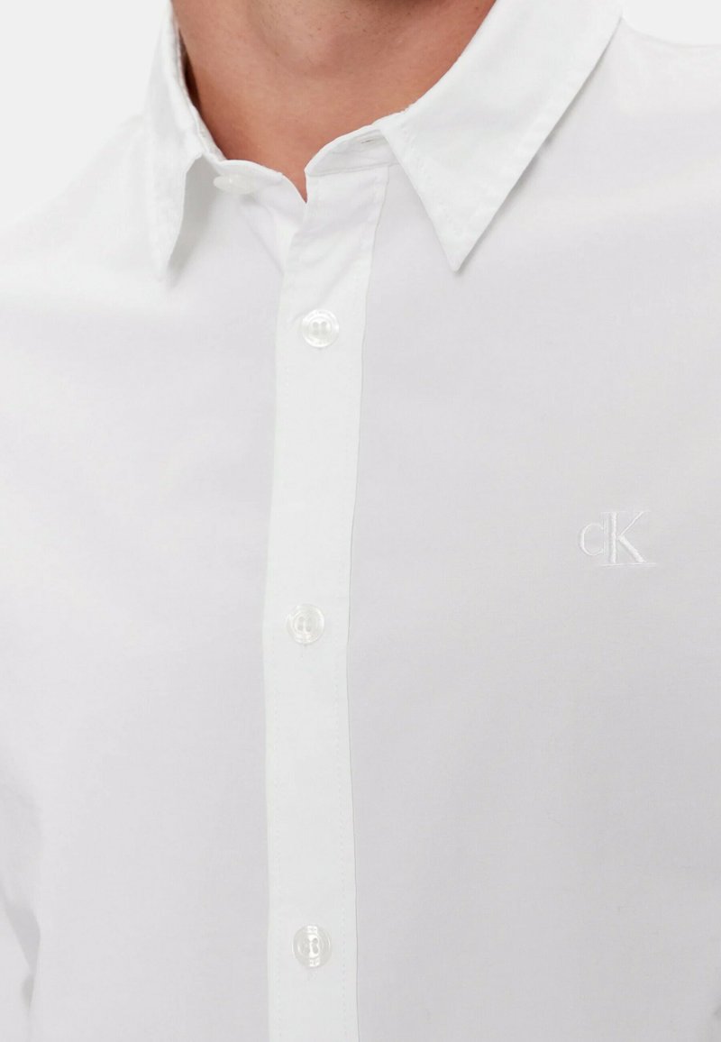 Biała koszula z guzikami, z wyraźnym kołnierzykiem, przezroczystymi plastikowymi guzikami i małym haftowanym logo. Gładki materiał i dopasowany krój.