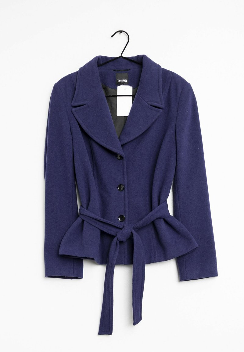Blazer bleu marine pour femme avec larges revers, trois boutons noirs, manches longues et ceinture en tissu nouée à la taille, suspendu sur un cintre noir.