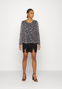 Top peplum preto com estampado floral, combinado com uma saia mini preta com fendas laterais. Modelo usa sandálias de salto pretas, em pé sobre um fundo branco.