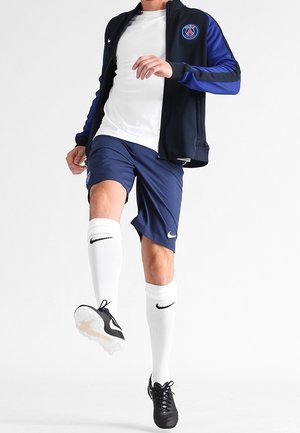 Męski piłkarz ubrany w granatowe spodenki, białe skarpetki i czarno-niebieską kurtkę, kopiący czarno-białą piłkę nożną.