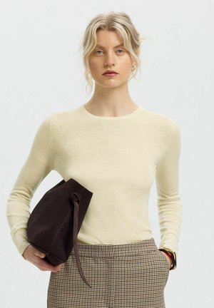 Kvinde med blondt hår iført cremefarvet langærmet top og ternede bukser, holder mørkebrun ruskindstaske, står mod en lys baggrund.