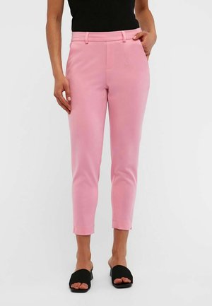 Femme portant un pantalon rose slim aux chevilles et des talons noirs ouverts, debout avec une main dans la poche.