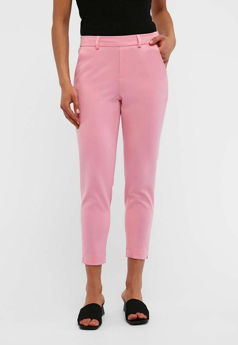Femme portant un pantalon rose slim aux chevilles et des talons noirs ouverts, debout avec une main dans la poche.