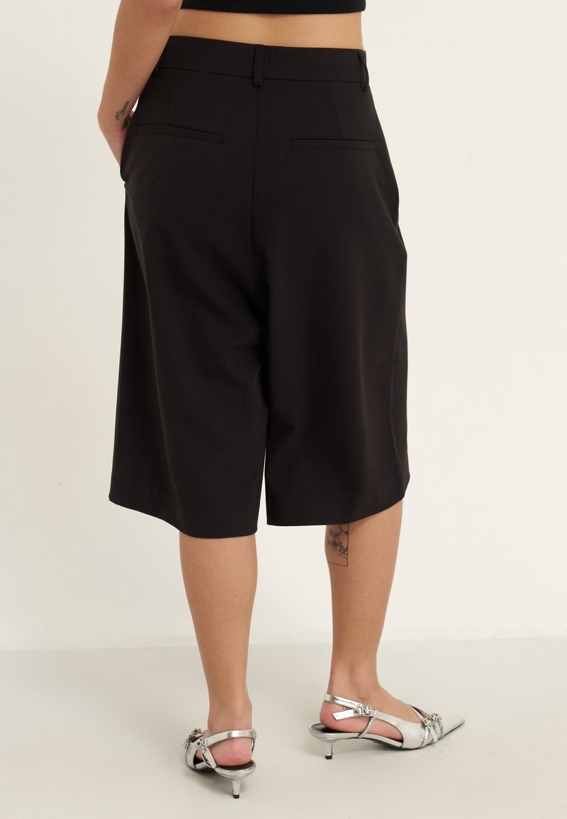 Personne portant un short noir sur mesure jusqu'aux genoux et des chaussures slingback argentées à talons bas, debout devant un fond clair uni.