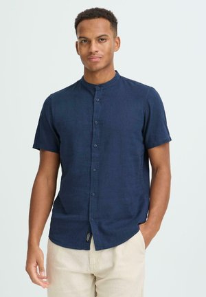 Junger Mann, der ein kurzärmeliges, marineblaues Hemd mit Stehkragen und helle beige Hose vor einem schlichten Hintergrund trägt.