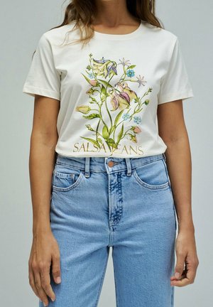 Vrouw die een wit T-shirt draagt met een kleurrijke bloemenprint en tekst "SALSA JEANS", gecombineerd met een high-waisted lichtblauwe spijkerbroek.