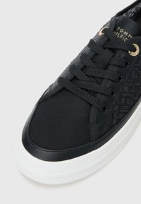 Svart sneakers med et teksturert mønster på siden, svarte snøringer, gulløyne og en hvit gummisåle. Laget av lerret og syntetiske materialer.