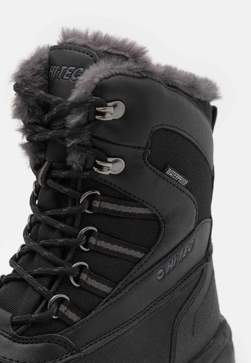 Botín negro impermeable con borde de piel de pelo, parte superior de cuero texturizado y tejido, ojetes de metal y cordones grises. Presenta logotipo y diseño reforzado.