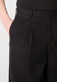 Pantalons en coton noirs avec des plis, dotés d'une poche latérale et d'une fermeture à bouton. Texture lisse et coupe cintrée.