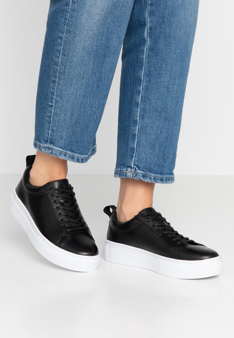 Personne portant un jean court bleu et des baskets en cuir noir avec des semelles blanches épaisses, debout sur un sol lisse et clair.
