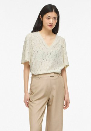 Kobieta w krótkim kremowym bluzce z teksturą i dekoltem w serek, włożona w beżowe spodnie z wysokim stanem, stojąca na jednolitym tle.