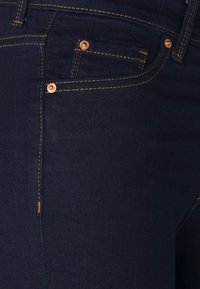 Tmavě modré džínové kalhoty, detail přední kapsy, malé mince kapsy, oranžové stehy a měděné nýty.