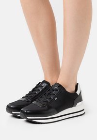 Baskets basses noires et blanches avec lacets, portées aux pieds, avec le logo de la marque sur le côté et un design de semelle rayée.