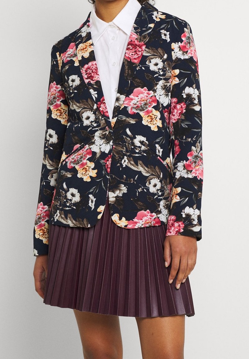 Femme portant un blazer bleu marine à motifs floraux par-dessus une chemise blanche boutonnée, associée à une jupe plissée bordeaux, debout devant un fond uni.