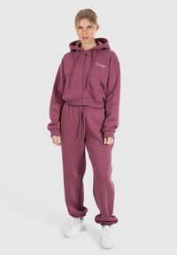 Felpa corta color bordeaux con zipper frontale e tasche, abbinata a pantaloni jogger coordinati, entrambi realizzati in tessuto morbido e testurizzato. Sneakers bianche.