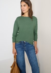 Maglione verde lavorato a maglia con scollo a barca, abbinato a jeans larghi blu. La modella tiene una borsa in suede marrone. Texture semplice e liscia.