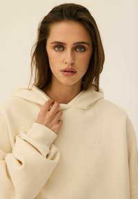 Kremowa bluza z kapturem wykonana z miękkiego materiału, z kieszenią typu kangur, ściągaczami przy mankietach oraz wytłoczonym logo na piersi.