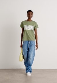 Grünes Baumwoll-T-Shirt mit fettem weißen Text, kombiniert mit hellblauen weiten Jeans und einer kleinen gelben Handtasche. Weiße Sneaker runden den Look ab.