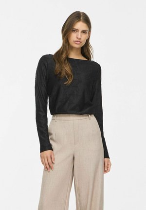 Une femme porte un haut noir texturé à manches longues et un pantalon beige à jambes larges, sur un fond de studio blanc uni.