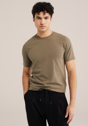 Tânăr cu păr creț purtând un tricou kaki cu mânecă scurtă și pantaloni negri, pozând pe un fundal bej simplu.