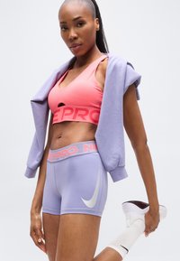 Ροζ αθλητικό σουτιέν με άνοιγμα, γκρι λογότυπο Nike σε λιλά σορτς με τη φράση 'NIKE PRO'. Το μοντέλο φοράει ανοιχτό μοβ φούτερ με κουκούλα και λευκά παπούτσια.