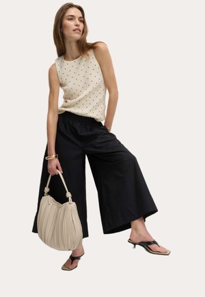 Top senza maniche color crema con pois neri, pantaloni cropped larghi neri, borsa beige plissettata e infradito nere con tacco basso.