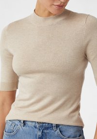 Beżowy sweter z krótkim rękawem, wykonany na drutach, z wysokim dekoltem. Posiada miękką fakturę i ściągacze, noszony z niebieskimi dżinsami.
