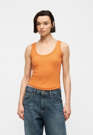 Femme aux cheveux courts et foncés portant un haut orange sans manches et un jean bleu, debout devant un fond clair uni.