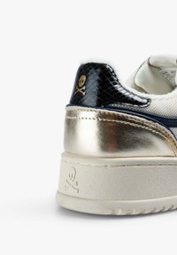 Zapatilla con tacón en texturas de serpiente en negro y dorado metálico, con un logo de calavera y huesos cruzados en la parte superior y en la suela.