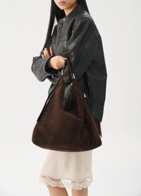 Frau in einer schwarzen Lederjacke und creamfarbenem Rock, die eine große braune Wildlederhandtasche vor einem schlichten weißen Hintergrund hält.