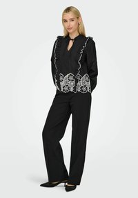 Blouse noire avec col volanté, décolleté en forme de clé et détails en dentelle florale blanche à l'ourlet. Associée à un pantalon noir et des talons à motifs.