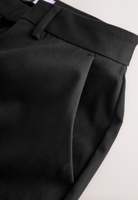 Gros plan sur un pantalon en tissu noir montrant la ceinture, les passants de ceinture et une poche latérale avec des détails de couture visibles sur un fond clair.